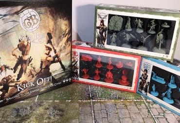 Photo showing several boxes of miniatures for the game Guild Ball on a table with the gameboard bellow. 