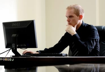 photo showing a man sat looking at a computer screen with a hand thoughtfully on his chin. 